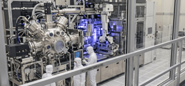Person observing three technicians in protective suits operating complex semiconductor machinery in a cleanroom