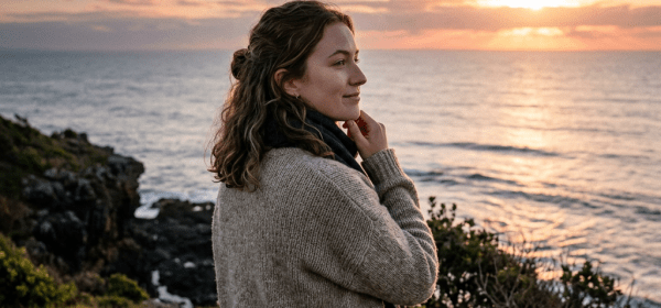 Woman standing on rocky coastline looking at ocean sunset
